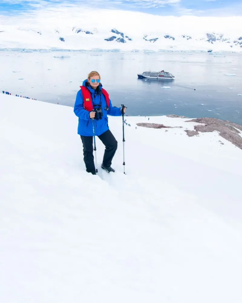 Eine Reisende mit Wanderstöcken auf einem Schneehang in der Antarktis, unten in der Bucht liegt ein Expeditionsschiff