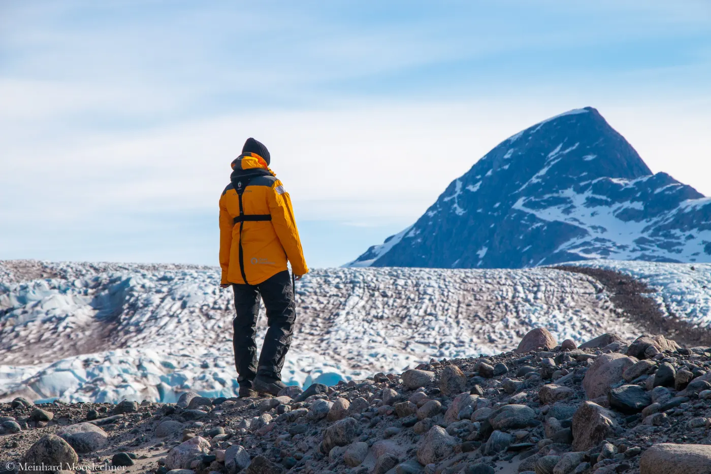 Eine Reisende in orangefarbener Expeditionsjacke steht auf einem Gletscher in Grönland und blickt zu einem Berg