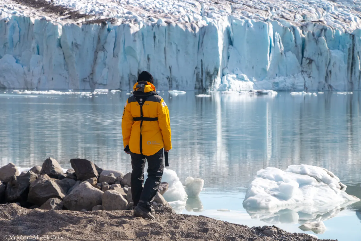 Eine Reisende in orangefarbener Expeditionsjacke steht am Ufer vor einer Gletscherkante in Grönland
