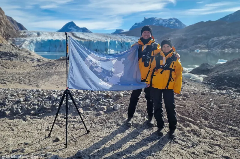 Meinhard Mooslechner und Daria Startseva in Expeditionsjacken mit einer Flagge vor einem Gletscher in Grönland
